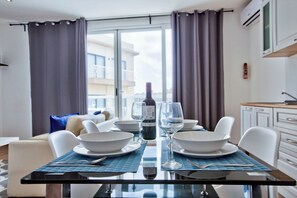 Ein Esstisch ist gedeckt mit weißen Schüsseln, Weingläsern und einer Weinflasche, mit Blick auf einen Balkon und ein Gebäude im Hintergrund.