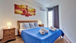 A bedroom with a bed, a nightstand, a lamp, a painting, and a window with curtains. - Central Sunny Apartment + Views of Valletta City (Sliema)