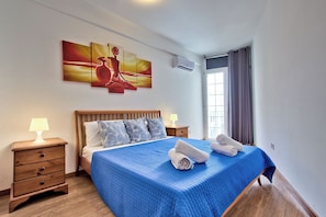 Ein Schlafzimmer mit einem Bett, einem Nachttisch, einer Lampe, einem Gemälde und einem Fenster mit Vorhängen.
