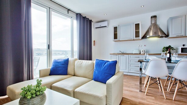 A modern living room with a sofa, a dining table, and chairs. There is a kitchen area with cabinets and a large window. - Central Sunny Apartment + Views of Valletta City (Sliema)