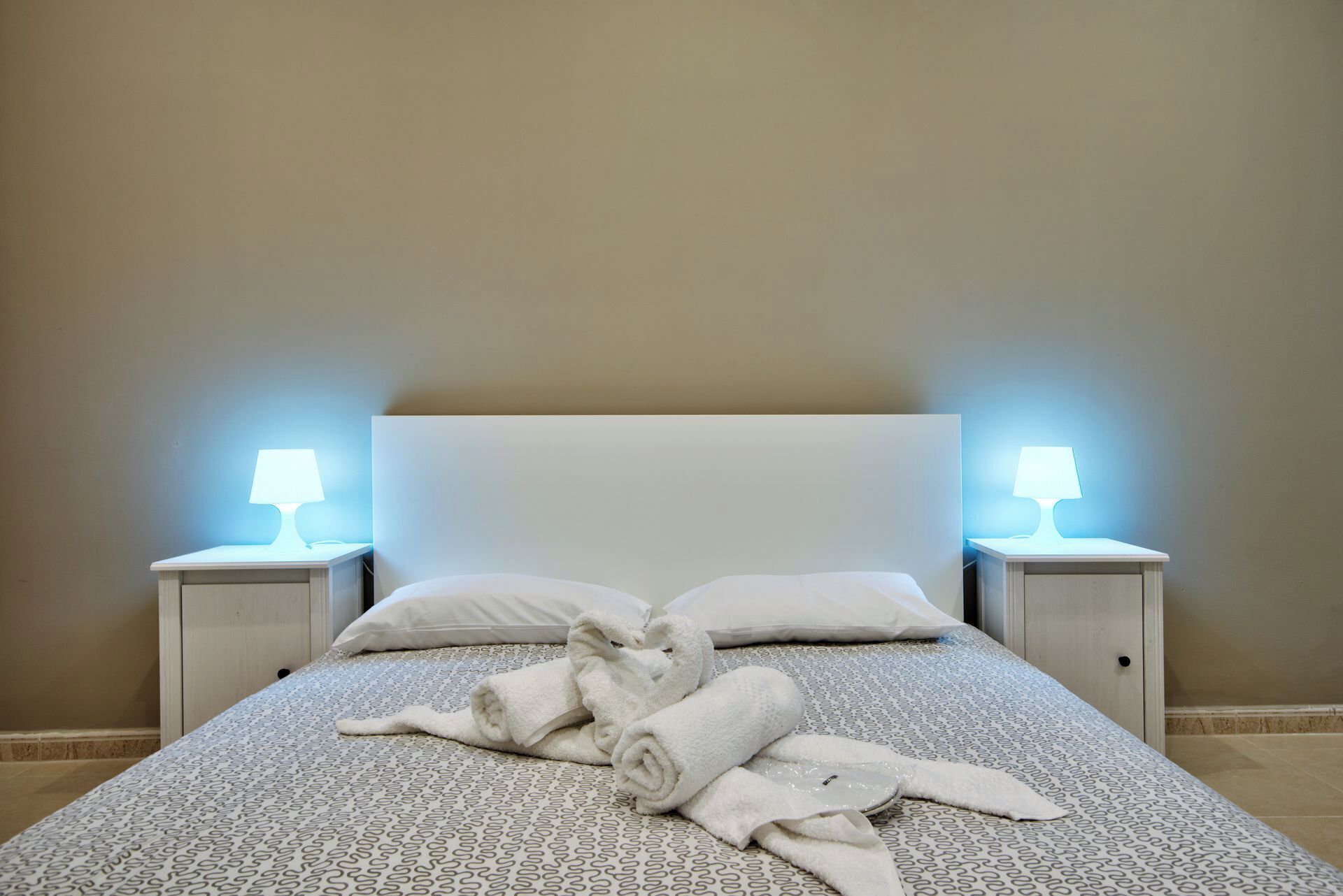 Un letto rifatto con cura, con una coperta a motivi, due lampade da comodino e asciugamani arrotolati sul letto.
