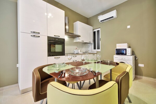 A compact kitchen with white cabinets, a built-in oven, a microwave, and a washing machine. There is a dining table with chairs and a glass top. - Central Flat With Traditional Maltese Balcony (Gzira)