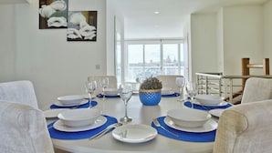 A dining room with a round table set for six, white plates, blue placemats, and clear glassware. A potted plant is on the table, and there are framed floral artworks on the wall. - Idyllic Penthouse Apartment With Sea View Balcony (Sliema)