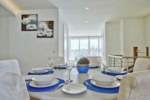 A dining room with a round table set for six, white plates, blue placemats, and clear glassware. A potted plant is on the table, and there are framed floral artworks on the wall. - Idyllic Penthouse Apartment With Sea View Balcony (Sliema)