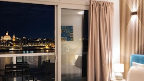A hotel room with a bed, a chair, a table, and a view of a cityscape at night. - Idyllic Penthouse Apartment With Sea View Balcony (Sliema)