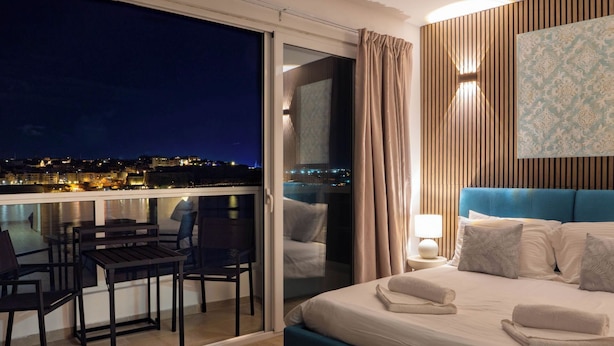 A modern hotel room with a balcony, a bed with blue headboard, and a city view at night. - Idyllic Penthouse Apartment With Sea View Balcony (Sliema)