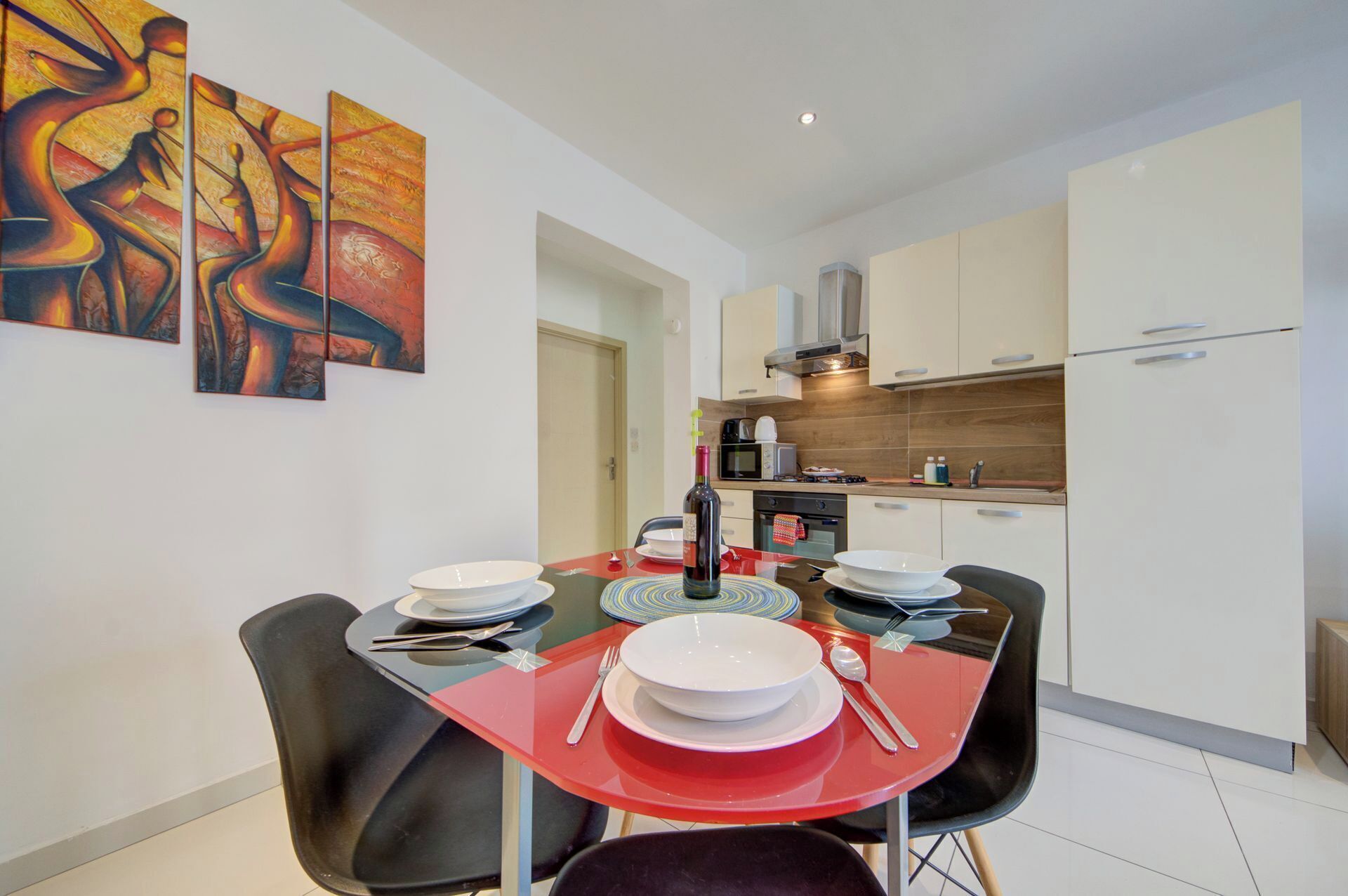 Une cuisine moderne avec des meubles blancs, un four à micro-ondes et une cuisinière. On y trouve une table à manger avec des chaises rouges et de l’assiette blanche.