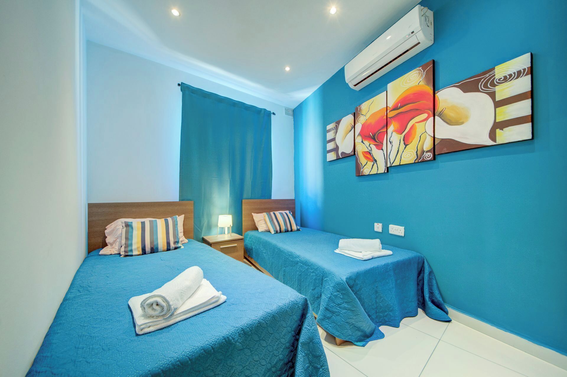 A hotel room with two beds, each with a blue bedspread and pillows, a nightstand with a lamp, and a wall with three paintings.