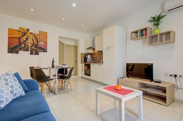 Een moderne woonkamer met een blauwe bank, een eethoek met stoelen, een keuken met een magnetron en een televisie op een houten dressoir.