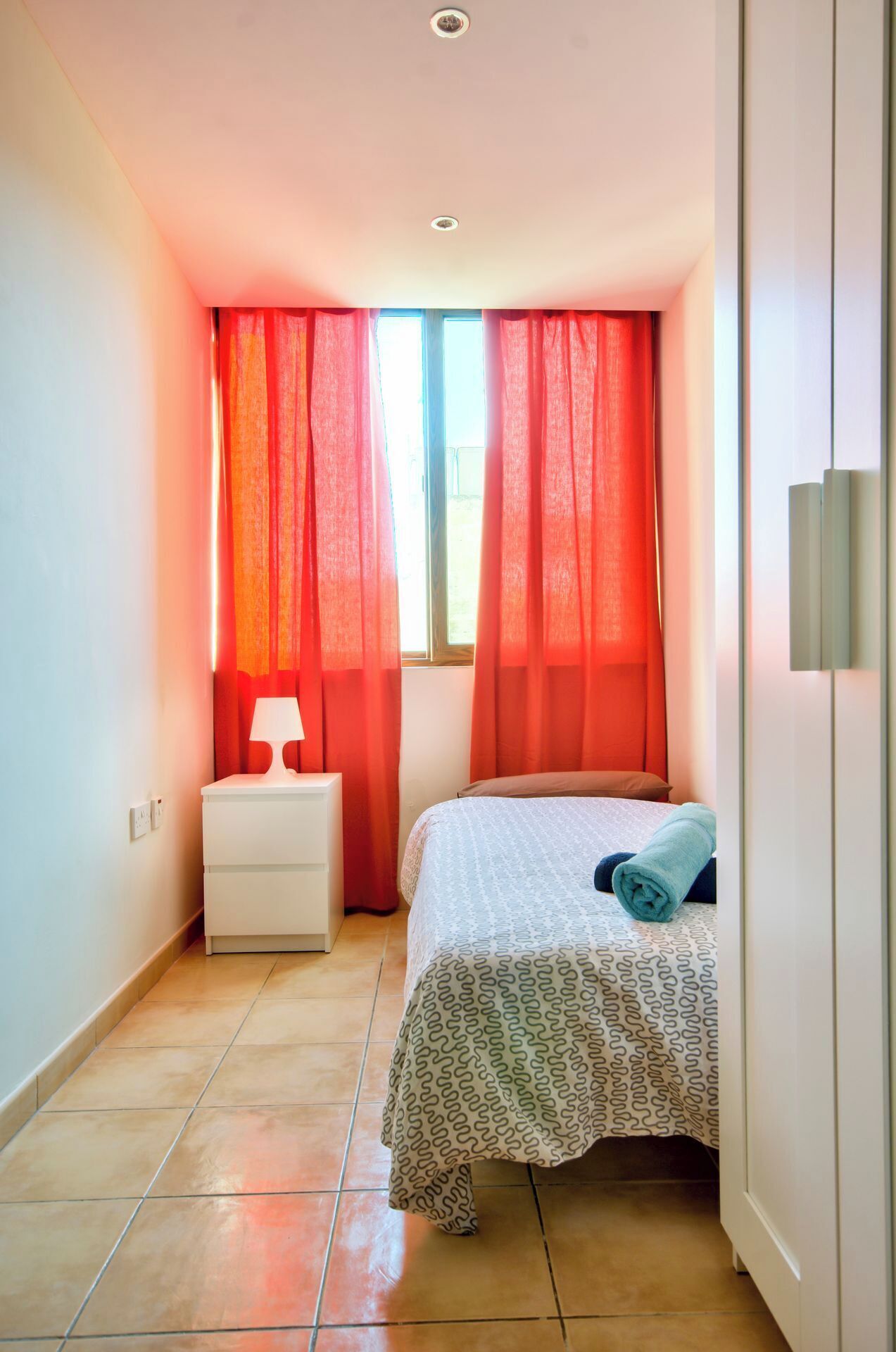 A small, well-lit bedroom with a bed, nightstand, and a window with red curtains.