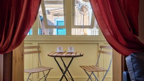 A room with a small table and two chairs, a window with curtains, and a view of buildings outside. - Delightful Apartment With Traditional Balcony (Sliema)