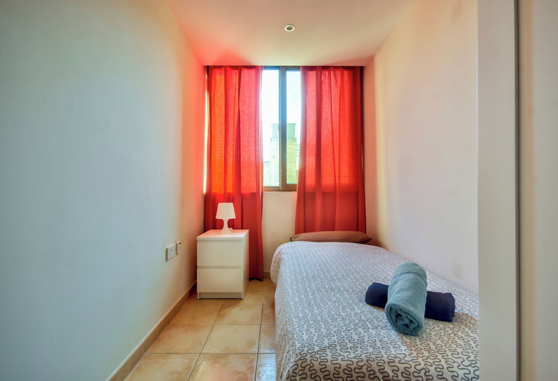 Une petite chambre d’hôtel étroite, avec un lit, une table de chevet et une fenêtre ornée de rideaux rouges.