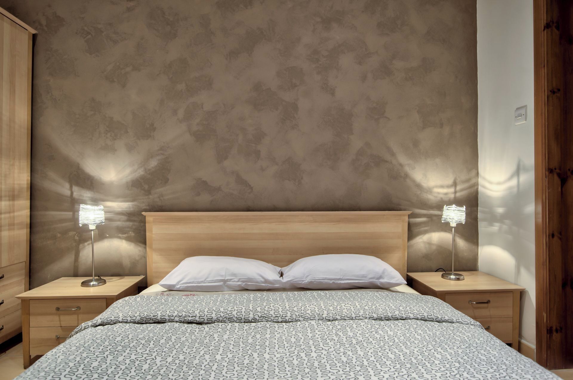 A bedroom with a wooden headboard, two bedside tables with lamps, a patterned bedspread, and a textured wall.