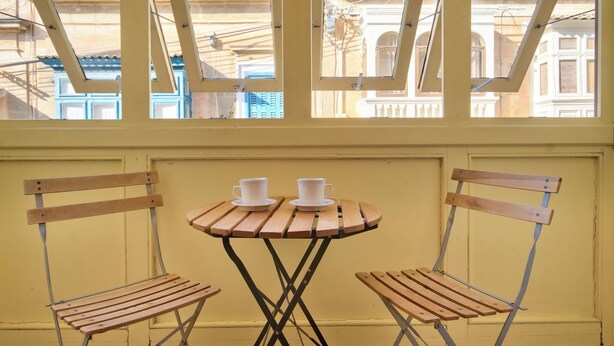 A small table with two chairs and two cups, set against a yellow wall with a window overlooking a building. - Delightful Apartment With Traditional Balcony (Sliema)