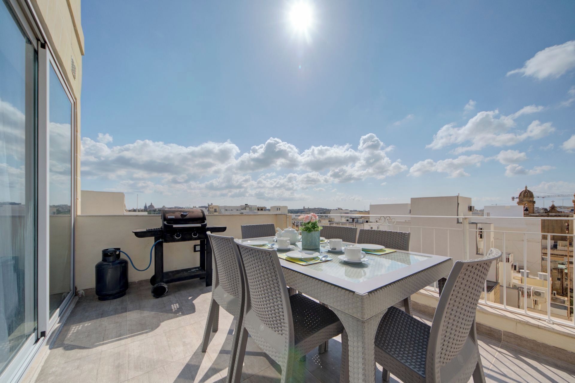 Un balcon avec une table dressée pour deux personnes, un barbecue et une vue sur le paysage urbain.