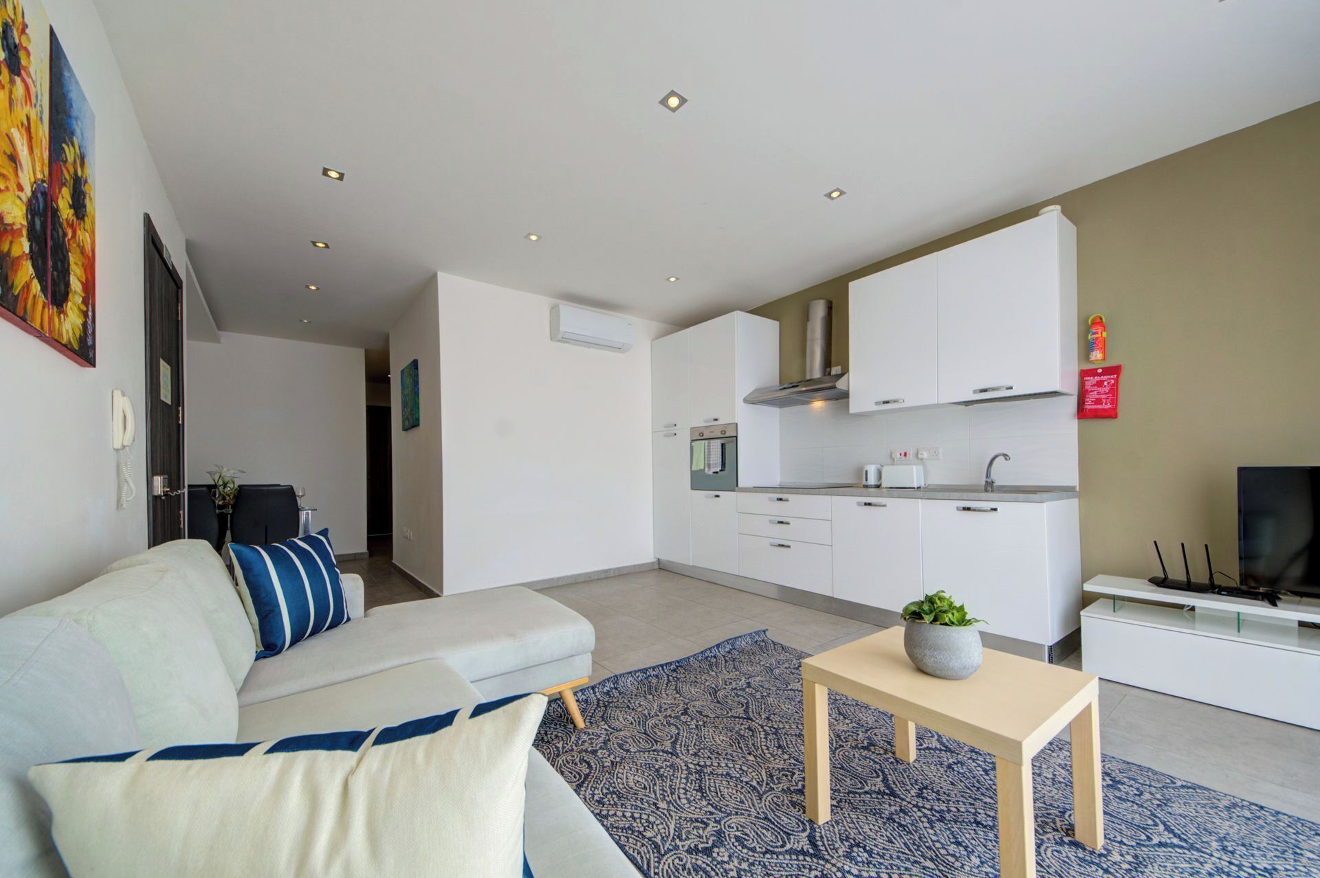 A modern living room with a sofa, a coffee table, and a TV. Adjacent is a kitchen with white cabinets, a stove, and a microwave.