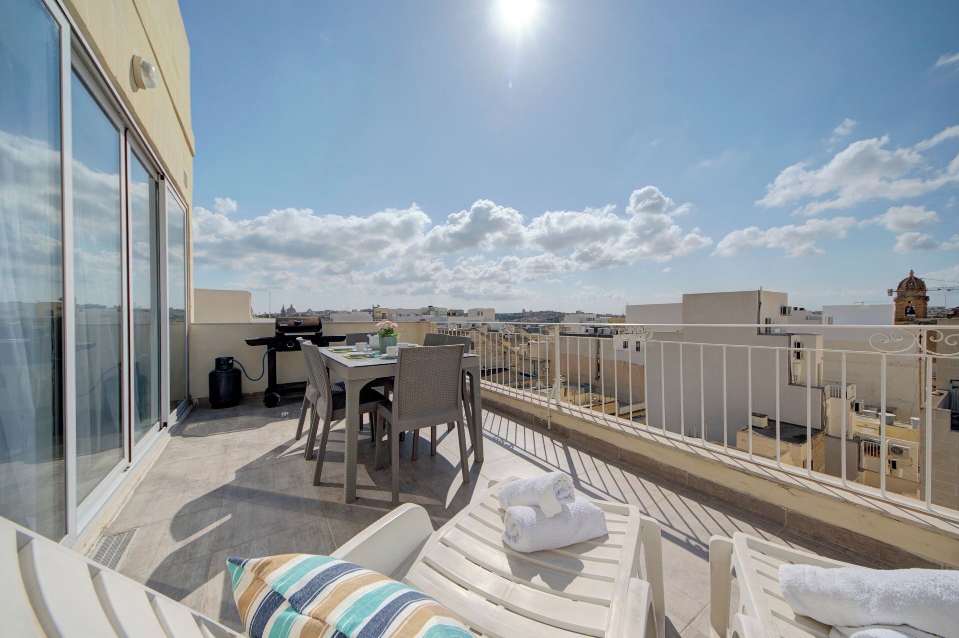 Eine Dachterrasse mit Tisch und Stühlen, einem Grill und Liegestühlen mit Handtüchern.