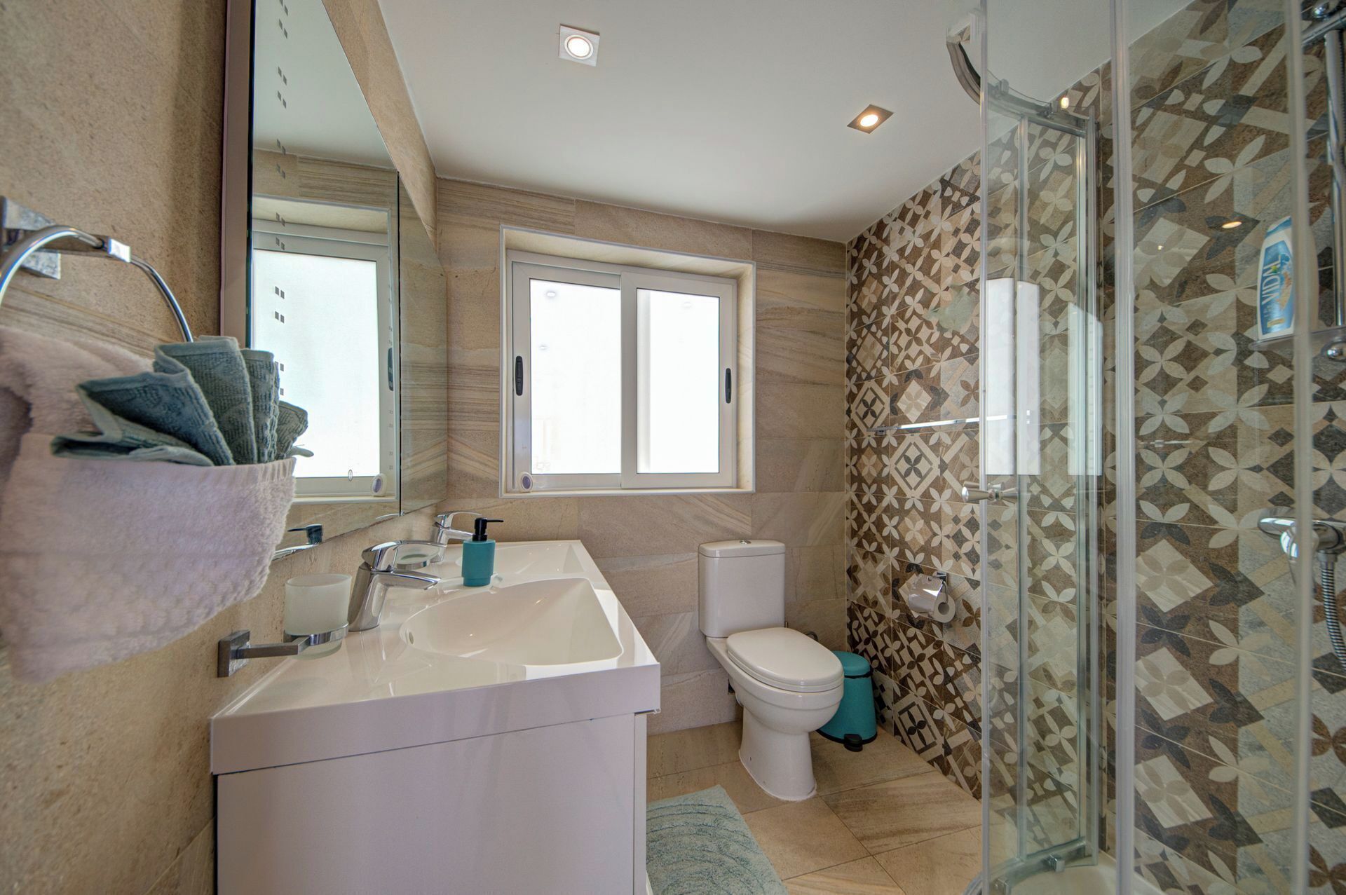 A bathroom with a white sink, a mirror, a window, a toilet, and a shower area with patterned tiles.