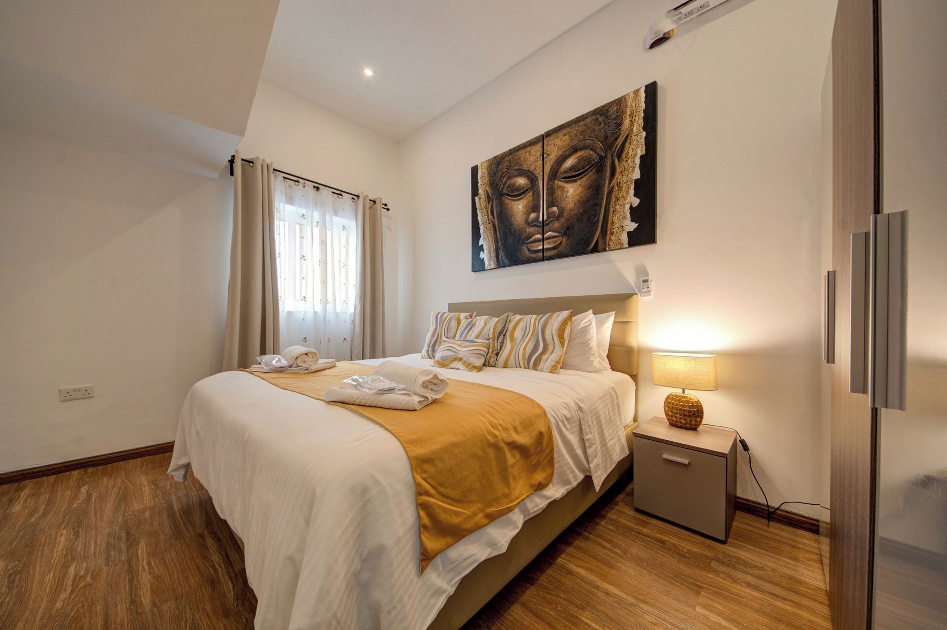 A hotel room with a large bed, a bedside table with a lamp, a wall-mounted artwork, and a small cabinet.