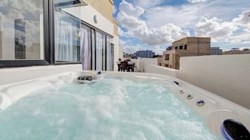 Uma banheira de hidromassagem no terraço, com cobertura de vidro, mesa e cadeiras, e vista para o horizonte da cidade ao fundo.