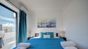 Ein Hotelzimmer mit einem großen Bett, zwei Nachttischen, einem Bild an der Wand und Blick auf Gebäude durch ein Fenster.