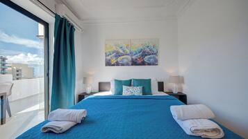 Quarto de hotel com uma cama grande, duas mesas de cabeceira, um quadro na parede e vista para prédios pela janela.