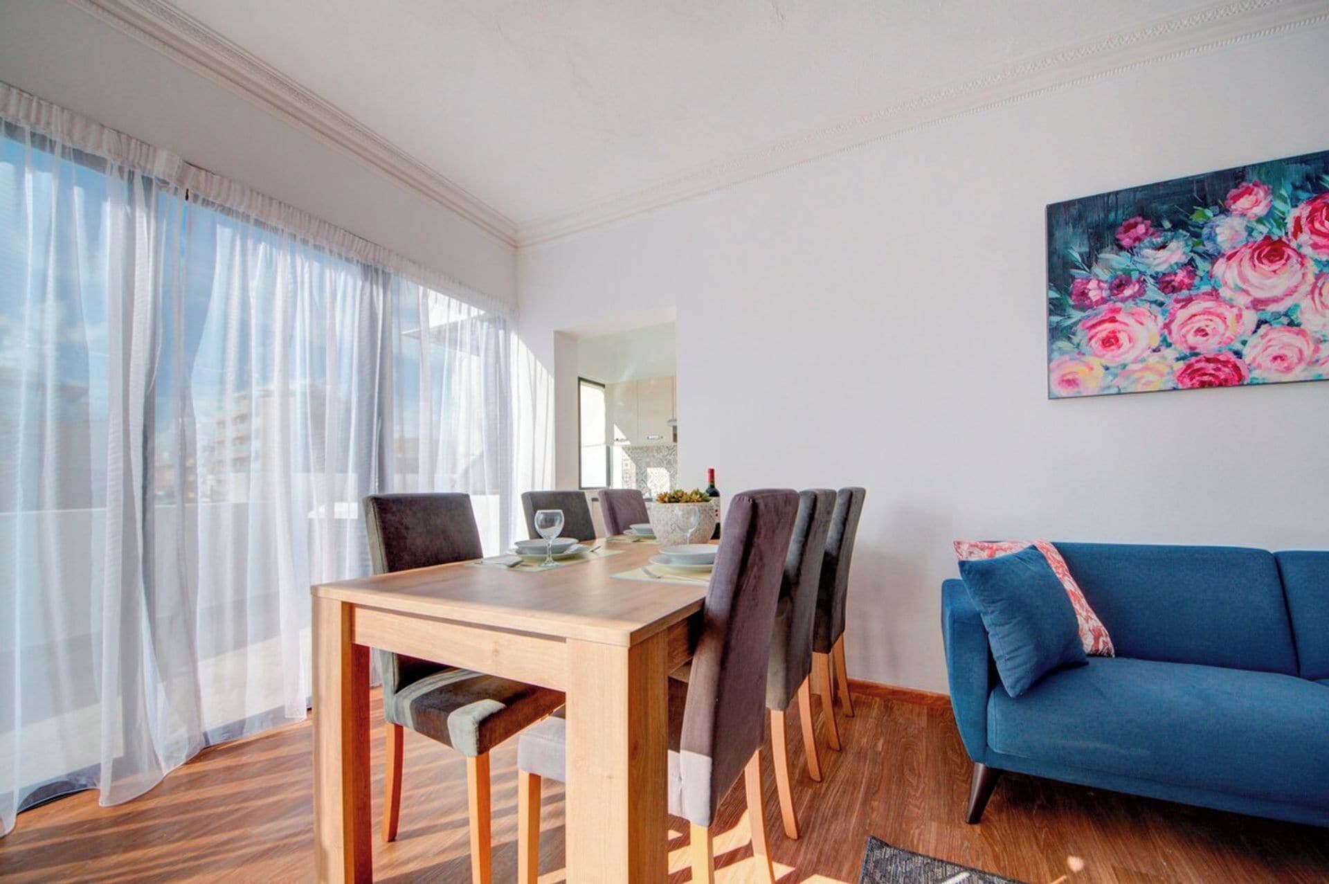 A dining area with a wooden table and chairs, a blue sofa, and a floral painting on the wall.