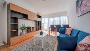 Ein modernes Wohnzimmer mit einem blauen Sofa, einem Couchtisch und eingebauten Holzregalen. Im Hintergrund befindet sich ein Essbereich mit Tisch und Stühlen.