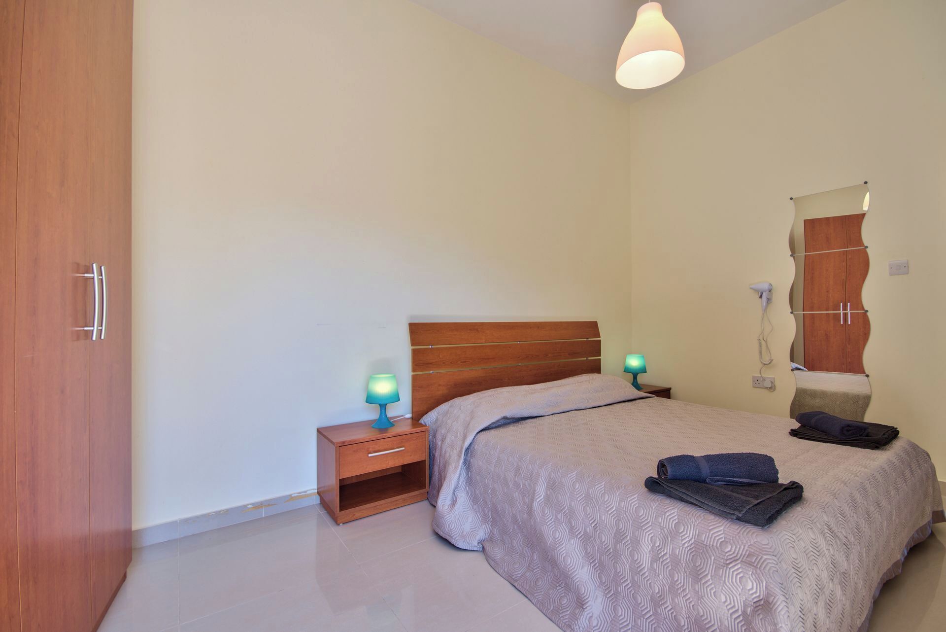 A bedroom with a single bed, a nightstand with a lamp, a wooden wardrobe, and a mirror on the wall.