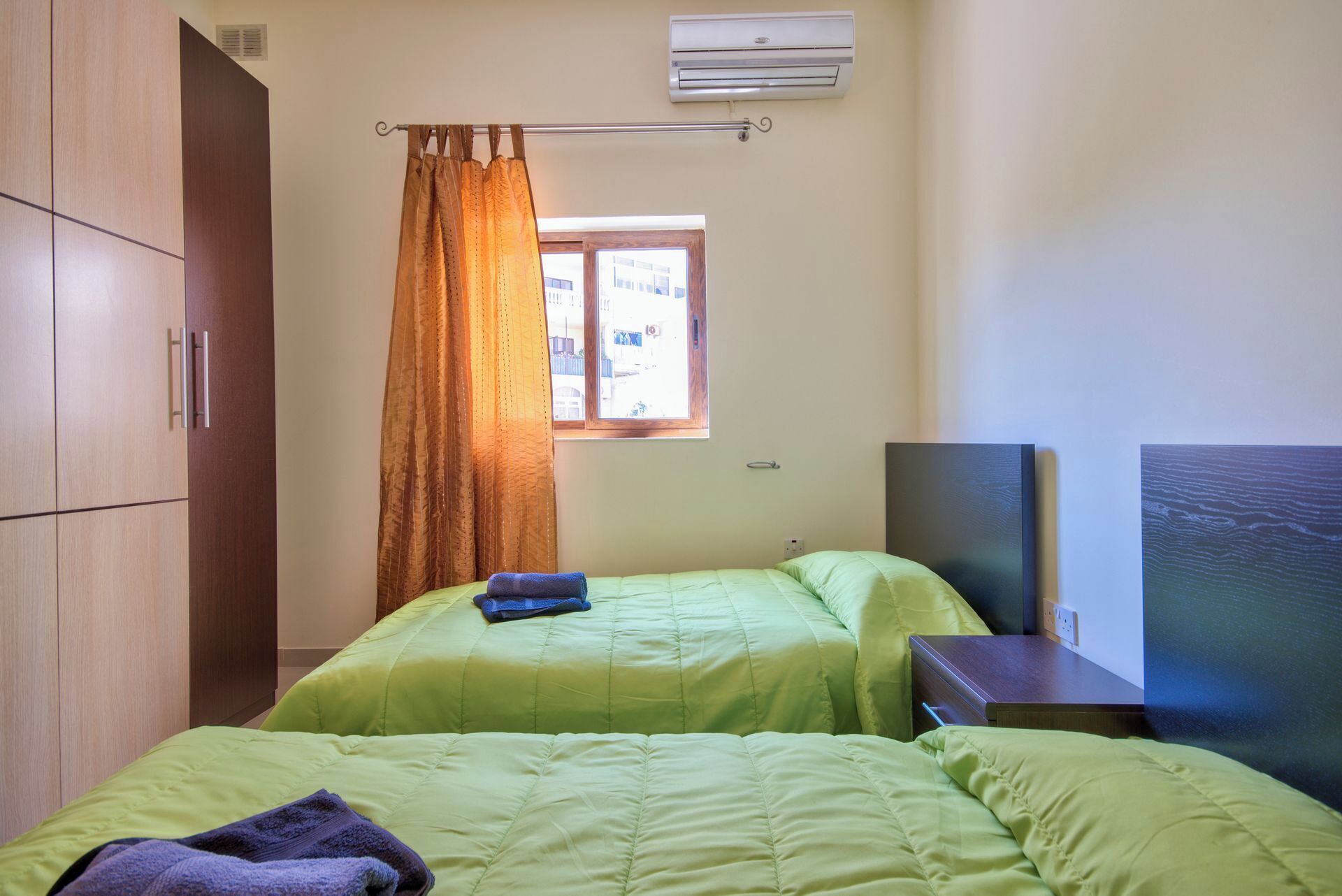 A room with two beds, a window with curtains, an air conditioner, and a wall-mounted TV.