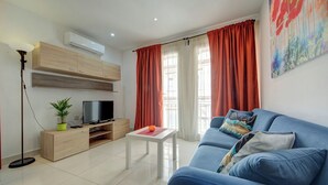 A modern living room with a blue sofa, a TV on a wooden cabinet, a small table with a plant, and red curtains. - Off the Ferries, Sliema, 2-bedroom Apartment (Sliema)
