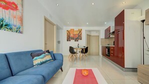 A modern living room with a blue sofa, a dining table with chairs, and a kitchen area with red cabinets. - Off the Ferries, Sliema, 2-bedroom Apartment (Sliema)