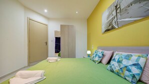 A hotel room with a green bedspread, a large bed with patterned pillows, a painting of a sailboat on the wall, and a door to the left. - Off the Ferries, Sliema, 2-bedroom Apartment (Sliema)