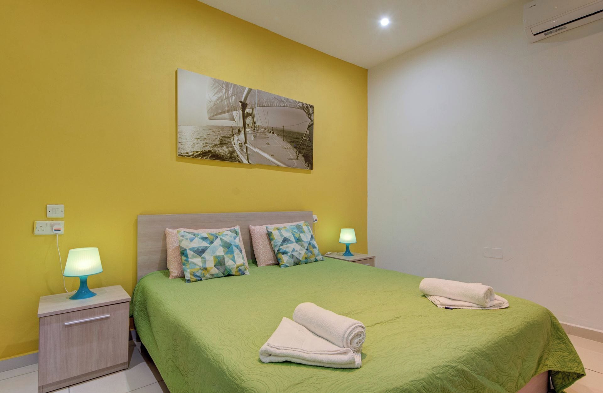 A hotel room with a green bed, two pillows, a painting on the wall, and two bedside lamps.