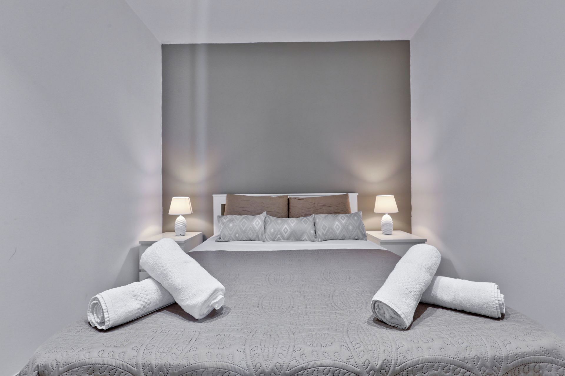 A modern bedroom with a large bed, two bedside lamps, and rolled towels on the bed.