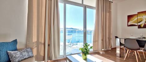 A modern living room with a sofa, a coffee table, and a dining area with a table and chairs. Large windows offer a view of the sea.