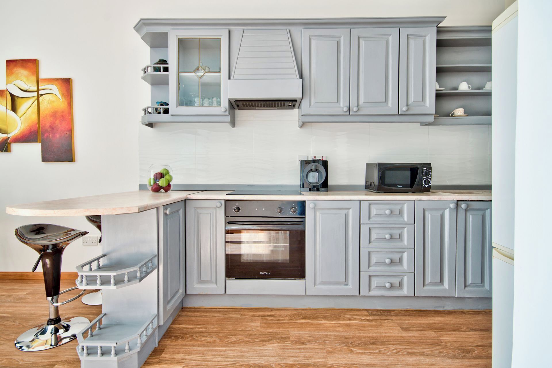 A modern kitchen with grey cabinetry, a built-in oven, a microwave, and a bar stool.