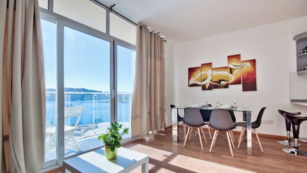 A modern dining area with a glass table, chairs, and a view of a body of water through large windows. - Stunning Home Overlooking Valletta City Skyline (Sliema)