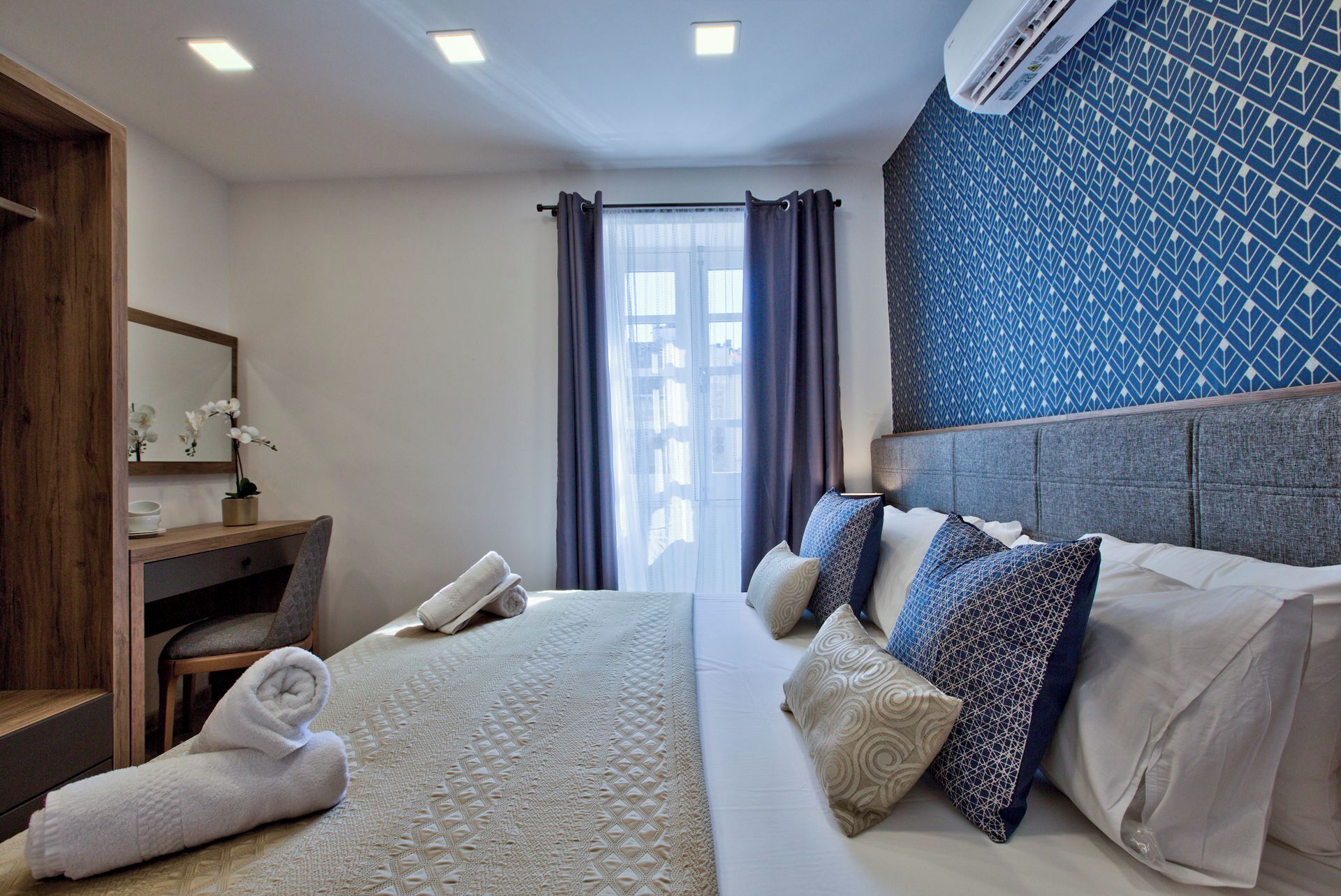 Habitación de hotel con cama, mesita de noche, espejo, ventana con cortinas y pared con diseño.