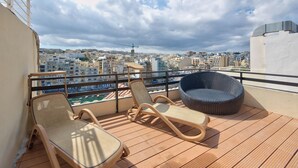 Ein Balkon mit zwei Liegestühlen und einem Korbsitzmöbel, mit Blick auf eine Stadtlandschaft, einschließlich Gebäude und einem entfernten Turm.