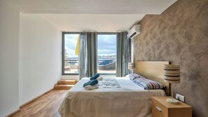Ein Schlafzimmer mit großem Fenster, einem Bett mit gestreiftem Kopfkissen, einer Nachttischlampe und einem Holznachttisch.
