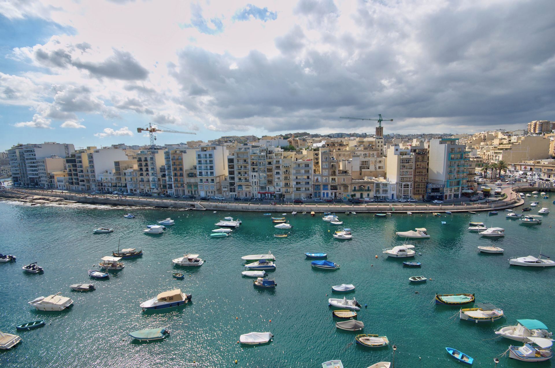 A coastal cityscape with a marina filled with boats, buildings with balconies, and a construction crane in the background.