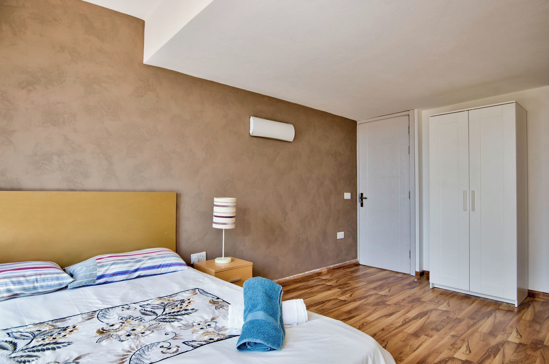 A bedroom with a bed, a nightstand, a lamp, a blue towel, and a wardrobe.