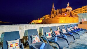 A seaside view with a row of lounge chairs with patterned cushions, a stone wall, and a historic building with a dome and spire in the background. - Enchanting Stone Villa With Pool and Jacuzzi (Valletta)