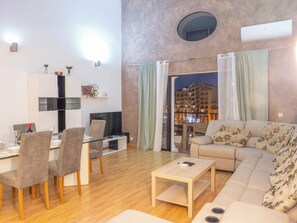 A modern living room with a sofa, coffee table, and dining area. There is a balcony with a view of buildings. - Stylish Modern Apartment With Spacious Living (St. Julian's)
