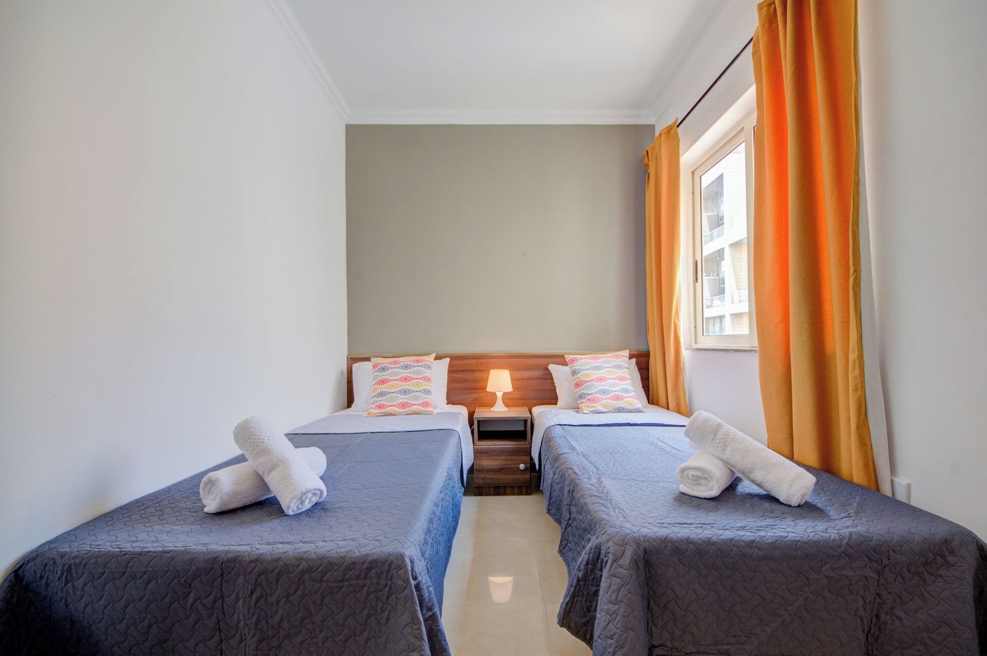 A small, clean room with two single beds, a nightstand with a lamp, and a window with curtains.