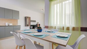 A modern dining area with a wooden table set for four, white chairs, and a view of a kitchen with grey cabinets and a green curtain. - Airy Top Floor Apartment With Stunning Terrace (St. Julian's)