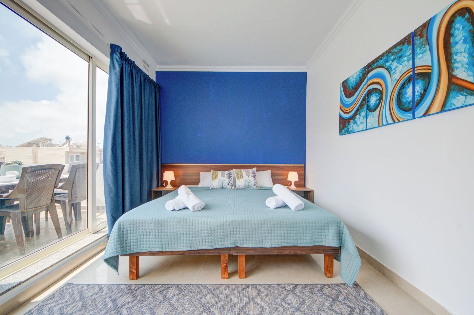 A hotel room with a large bed, a view of the city, and a blue wall.