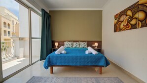 A hotel room with a large window, a bed with a blue bedspread, two pillows, a nightstand, and a painting on the wall. - Airy Top Floor Apartment With Stunning Terrace (St. Julian's)