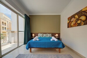 Ett hotellrum med ett stort fönster, en säng med ett blått överkast, två kuddar, ett nattduksbord och en tavla på väggen.